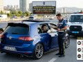 modifiyeli araç cezası denetimi: Trafik polisi, mavi bir Volkswagen Golf'ün cam filmini dijital cihazla kontrol ederken, arka kısımda diğer polis spoilerı inceliyor. Yan tarafta 'CAM FİLMİ %50 YASAL', 'SUNROOF İşlenmeli', 'SPOILER Abartı YASAK' yazılı bilgi paneli yer alıyor. Arka plan şehirlerarası yol ve trafik.