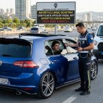 modifiyeli araç cezası denetimi: Trafik polisi, mavi bir Volkswagen Golf'ün cam filmini dijital cihazla kontrol ederken, arka kısımda diğer polis spoilerı inceliyor. Yan tarafta 'CAM FİLMİ %50 YASAL', 'SUNROOF İşlenmeli', 'SPOILER Abartı YASAK' yazılı bilgi paneli yer alıyor. Arka plan şehirlerarası yol ve trafik.